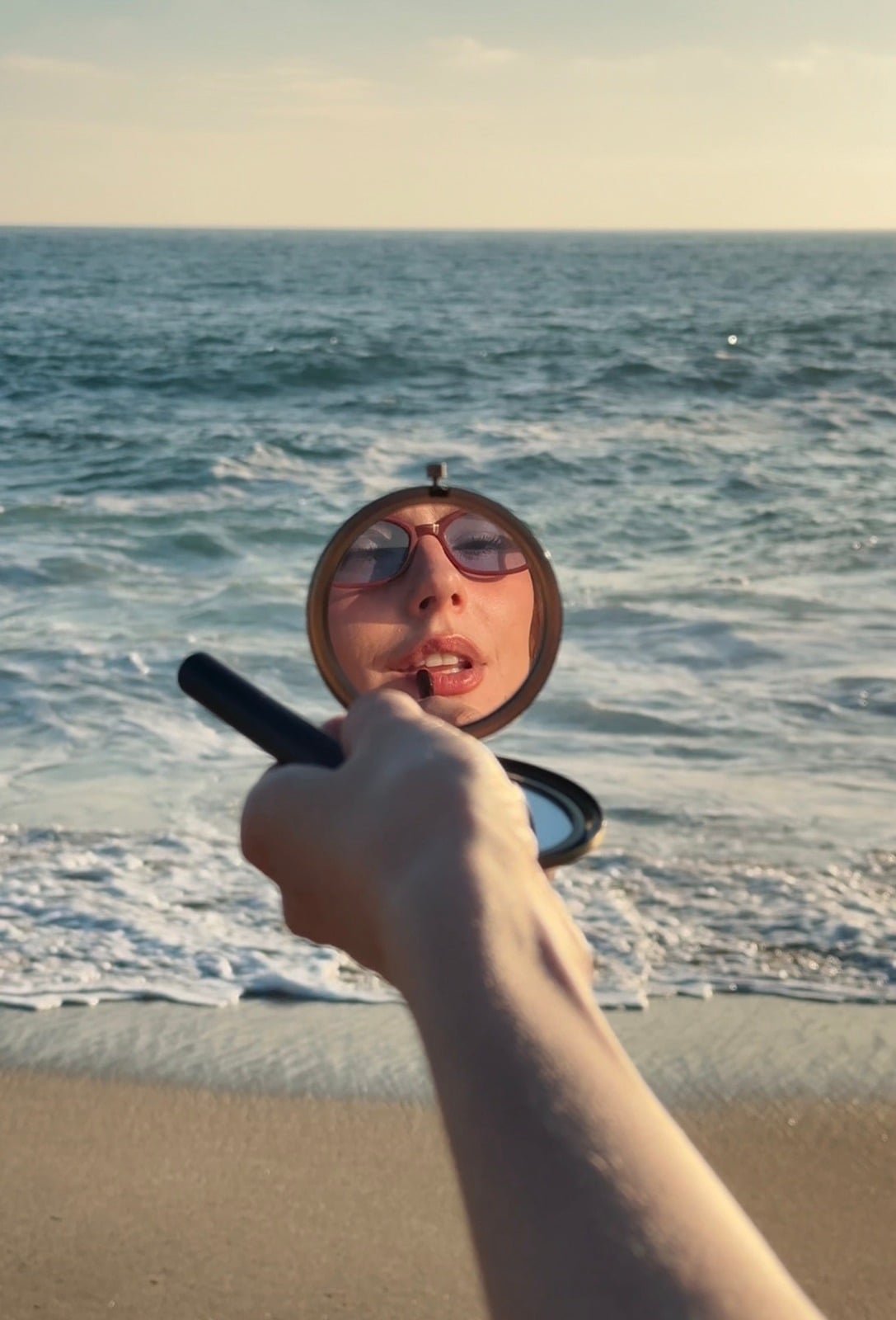 Compact mirror held up against the sea, reflecting a face in sunglasses