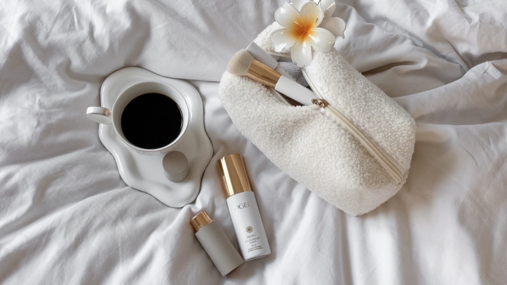 OGEE skincare primer beside a cup of coffee and a bouclé makeup bag on white sheets
